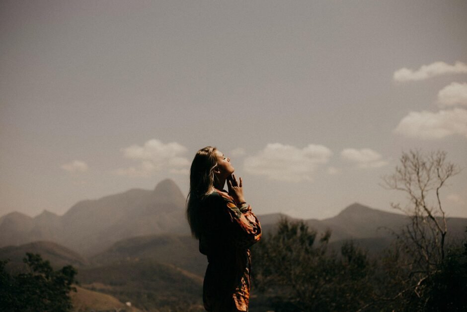 Silhouette of a person standing in profile against a mountainous landscape at sunset, head tilted back with eyes closed in a peaceful, meditative pose. The mountains are layered in the background under a cloudy sky, creating a serene, contemplative atmosphere.