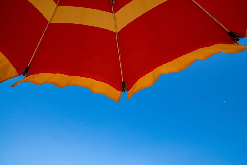 A bright red and orange striped beach umbrella against a vivid blue sky, photographed from below. The umbrella's pattern features bold alternating stripes, and its scalloped edges create a striking contrast against the clear summer sky.
