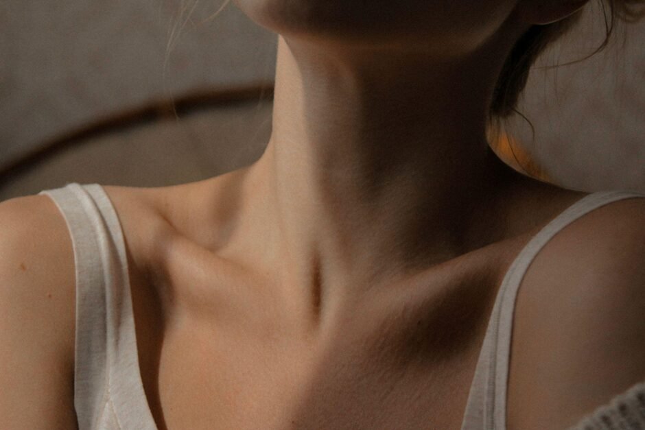 A close-up photograph of a person's neck and collarbone area, wearing a white tank top. The image is shot in warm, moody lighting with soft shadows, focusing on the natural contours and texture of the skin. The composition emphasizes the delicate anatomy of the neck and décolletage area, creating an artistic representation of skin texture and natural beauty.