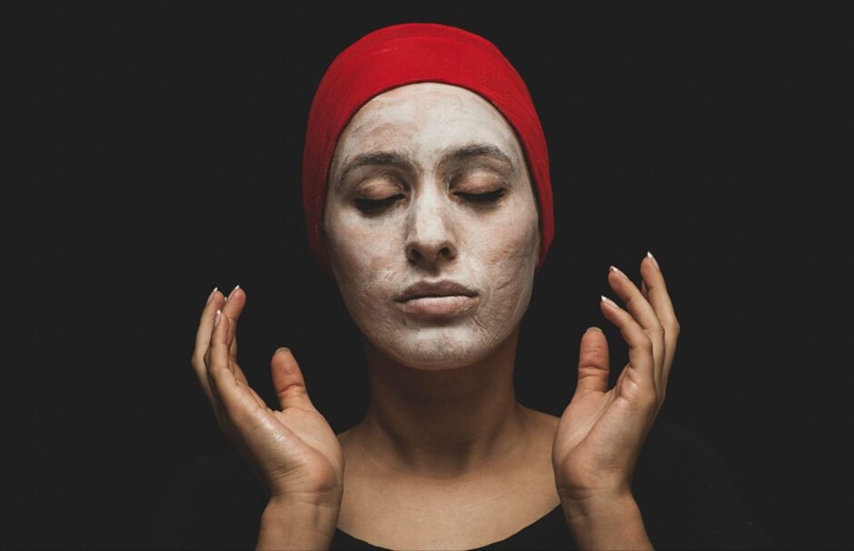 A close-up portrait of a person wearing a red beanie against a dark background. Their eyes are closed in a serene expression while wearing a face mask or treatment. Their hands are raised on either side of their face in a meditative pose. The image conveys skincare, self-care, and tranquility.