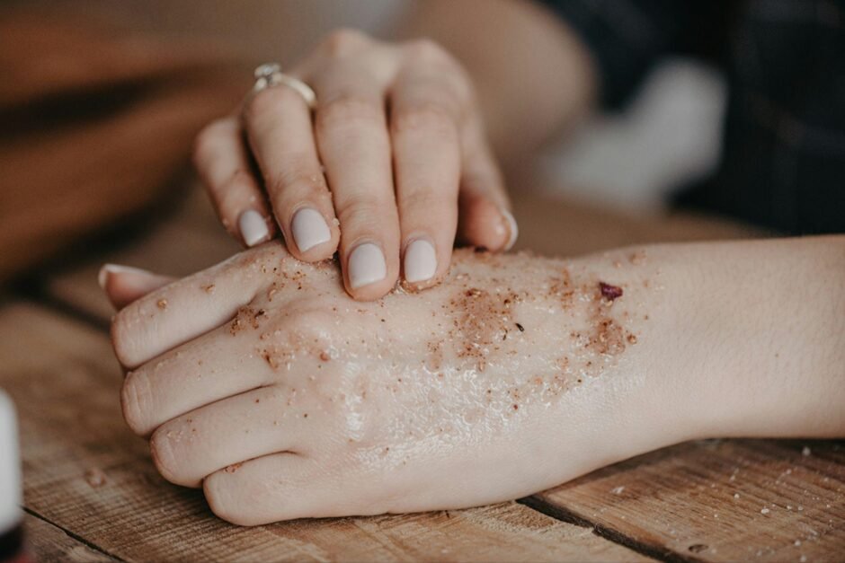 Close-up of hands gently applying a coffee or sugar scrub to pale skin. One hand wearing an engagement ring with neutral-toned manicure is seen exfoliating the other hand. The scene is captured on a wooden surface with a moody, natural lighting effect that emphasizes the texture of both the scrub and skin.