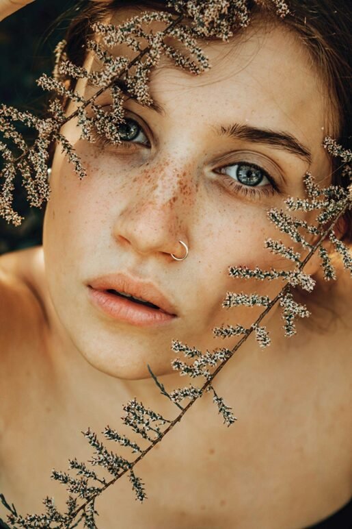 A close-up portrait in warm golden light shows a person with freckles, bright blue eyes, and a nose piercing. Delicate dried flowers or plant stems with tiny white buds create an artistic overlay across the image, casting gentle shadows on their face and creating a dreamy, natural aesthetic.