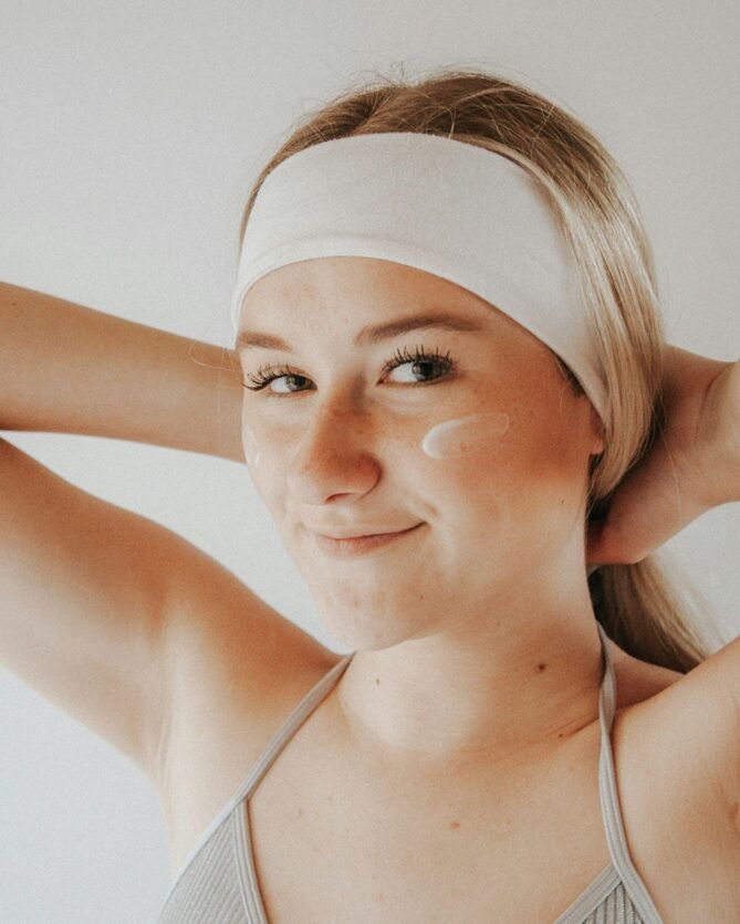 A close-up portrait of a person wearing a white headband and gray tank top. They have blonde hair pulled back and are smiling warmly while applying skincare product, shown by a small dab of cream on their cheek. The image has a soft, warm filter and shows natural, glowing skin against a plain white background.