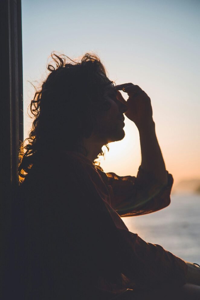 A silhouette of a person with curly hair seen in profile against a sunset sky, touching their face thoughtfully while backlit by warm golden light. Soft natural lighting creates a contemplative mood.