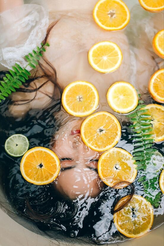 Fresh orange slices and fern leaves floating in clear water with ice cubes, creating a refreshing and natural spa-like setting. The vibrant citrus circles contrast beautifully with the delicate green ferns, suggesting skin rejuvenation and vitamin C benefits.